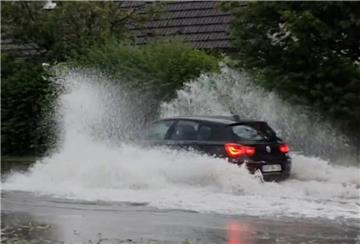 Ein Auto fährt durch das stehende Wasser auf einer Straße im Ort und schiebt eine Bugwelle vor sich her.