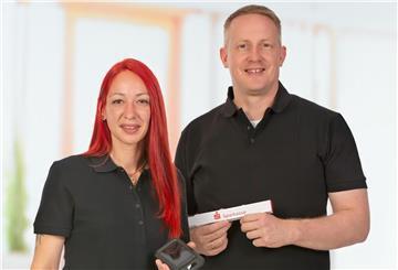 Zwei Energieberater - eine rothaarige Frau und ein blonder Mann in schwarzen Shirts - lächeln in die Kamera und halten ein Sparkasse-Schild in der Hand.