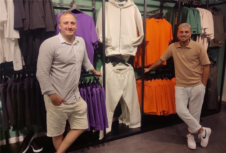 Nachhaltige Mode: Fast Fashion war gestern Joschka Traue und Berc Mardiros stehen am Kleiderständer. Auf den Kleiderständern befinden sich Hoodies, T-Shirts und Jogging-Hosen in weiß, lila, orange und grün.