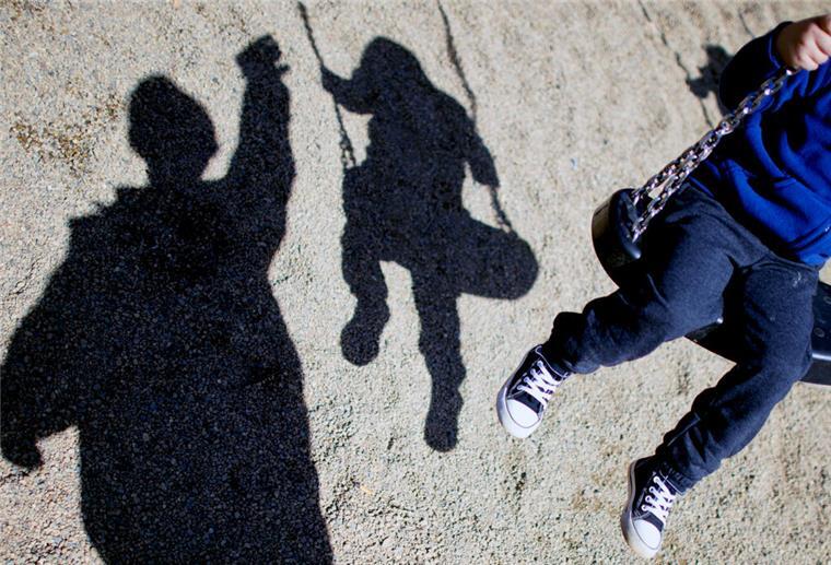 Eine Person steht vor einem Kind, das auf einer Schaukel sitzt. Im Hintergrund sind die Schatten zu erkennen. 