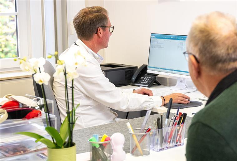 Ein älterer Herr sitzt im Raum und unterhält sich mit einem Arzt, der auf seinen Bildschirm guckt.  Kürzlich ist der Ruf nach einer neuen Praxisgebühr laut geworden – zur Eindämmung von Ärzte-Hopping und zur Entlastung der Kassen.