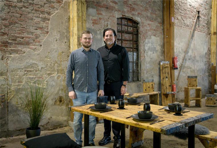 Zwei Männer stehen in einer Halle, in der Möbel und Geschirr stehen.
