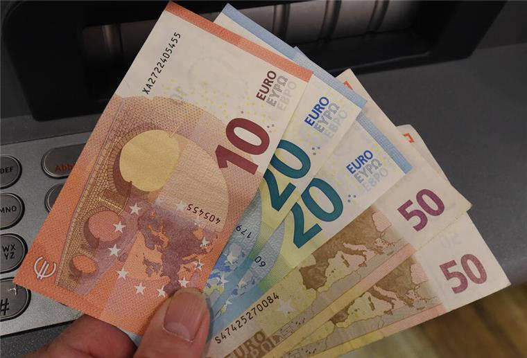 Eine Person hält Geldscheine vor einem Geldautomaten in die Kamera. Millionen Bürger bekommen im September einen Zuschuss. 