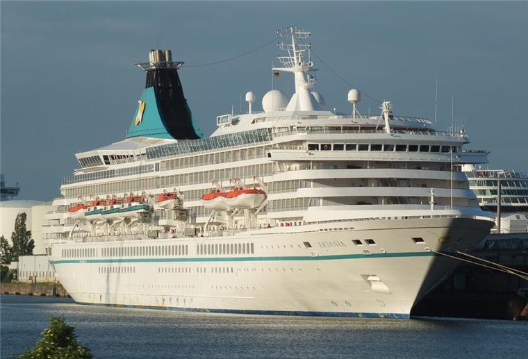 „Artania“ will zu einer Mega-Weltreise starten - Doch erst muss sie nach Bremerhaven Das Kreuzfahrtschiff „Artania“ liegt im Hafen. Das weiße Schiff mit türkisfarbenen Details verfügt über mehrere Decks, auf denen Rettungsboote zu sehen sind. Der markante Schornstein trägt das Logo von Phoenix Reisen.
