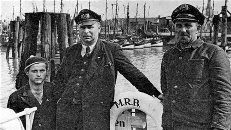Vor 70 Jahren: Die Seenotretter finden die im Herbststurm vermisste „Nossan“ Schwarz-Weiß-Aufnahme von drei Männern an Bord eines Schiffes im Hafen.