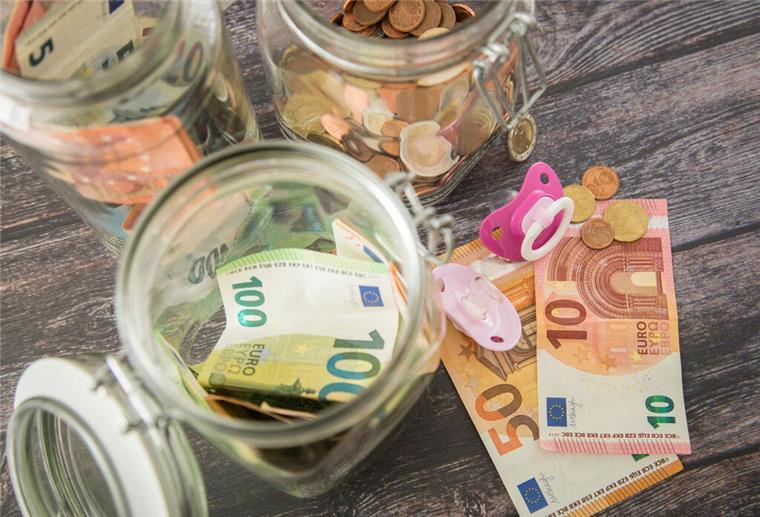 Bis zu 297 Euro im Monat! Dieser Zuschuss entlastet Familien mit Kindern Geldscheine und Münzen stecken in Gläsern, die auf einem Tisch stehen. Daneben liegen zwei Schnuller.