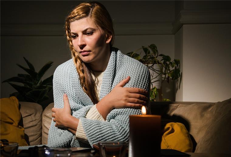 Eine Frau in einer Strickjacke schlingt offenbar frierend die Arme um ihren Oberkörper. Davor brennt eine Kerze.