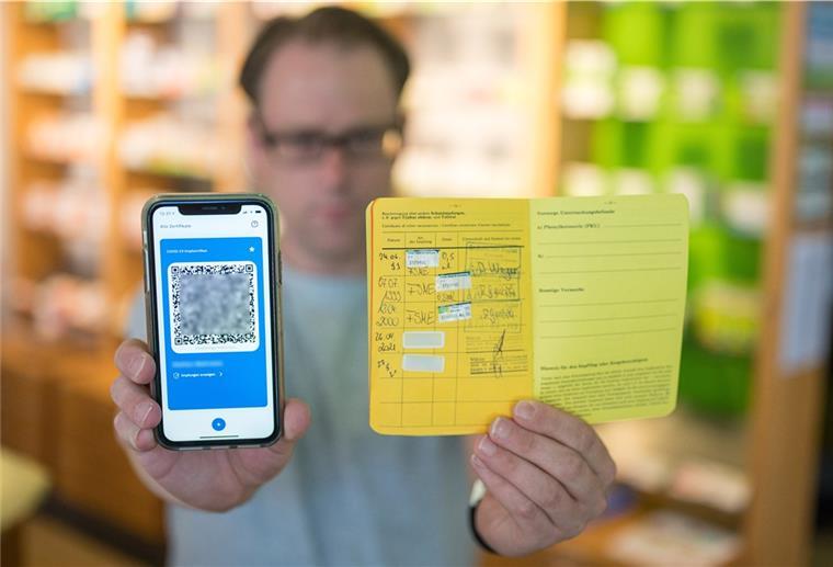 EU-Impfnachweise ohne Booster nur noch neun Monate gültig Apotheker Stefan Behrens hält ein Smartphone mit dem QR Code eines digitalen Impfausweises und ein Impfbuch in der Hand.