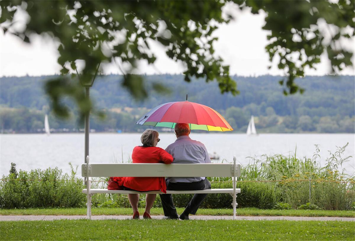 Ein Mann und eine Frau sitzen am Ufer eines Sees mit einem Sonnenschirm in der Hand auf einer Bank.