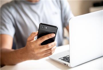 Ein Mann hält ein Handy in der Hand und sitzt vor dem Laptop