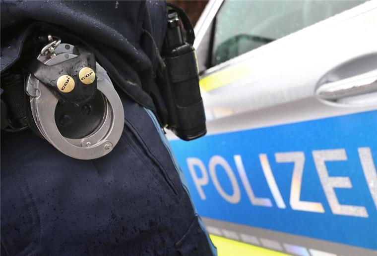 Im Vordergrund ein Paar Handschellen am Gürtel eines Polizisten, der vor einem Polizeiauto steht.