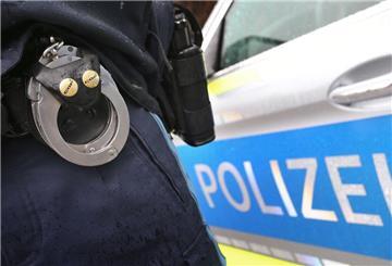 Im Vordergrund ein Paar Handschellen am Gürtel eines Polizisten, der vor einem Polizeiauto steht.
