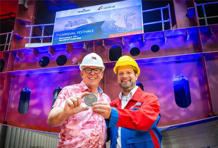 Meyer Werft legt neues Kreuzfahrtschiff für Carnival Cruise Line auf Kiel Zwei Männer in Schutzhelmen und Arbeitskleidung halten bei der Kiellegung der „Carnival Festivale“ eine Gedenkmedaille in den Händen. Im Hintergrund ist ein Schiffsrumpf mit einem Bildschirm zu sehen, der den Namen des Kreuzfahrtschiffes anzeigt.