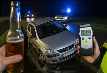 Ein Auto in einem Wassergraben, im Hintergrund Polizeifahrzeuge. Im Vordergrund ein Atenalkohol-Messgerät, dass die Zahl 2,38 anzeigt, und eine Flasche Bier in einer Hand.