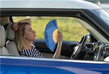 Eine Frau fächert sich im Auto frische Luft zu. Hitzefalle Auto: So bleiben Autofahrer cool. Schon nach wenigen Minuten kann ein Auto zur tödlichen Hitzefalle werden – vor allem für Kinder und Tiere. Immer zuerst lüften, nie bei direkter Sonne parken und auf reflektierende Abdeckungen achten.