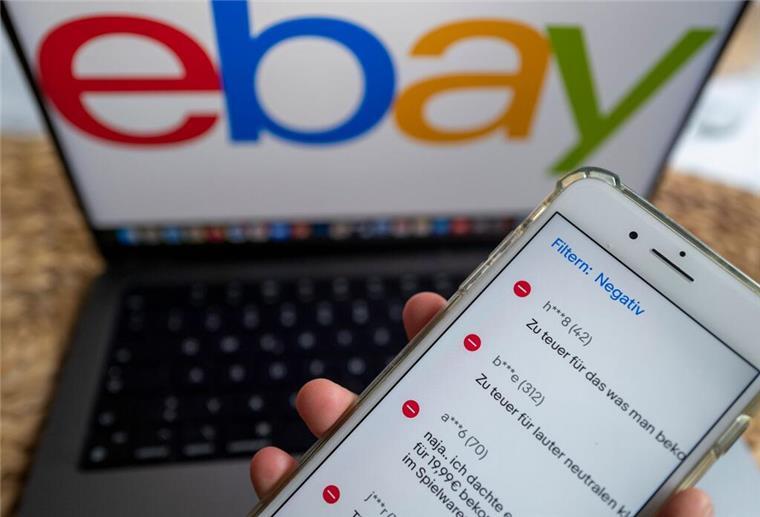 Eine Hand hält ein Handy. Dahinter ist ein Bildschirm mit dem Ebay-Schriftzug zu sehen.