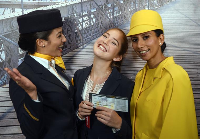 Casting für Flugbegleiter: Lufthansa geht neue Wege Sie hat das Casting bestanden: Joana Wollweber (Mitte) hält mit großer Freude neben zwei Flugbegleiterinnen, von denen eine (rechts) eine historische Uniform trägt, ihre erfolgreiche Casting-Teilnahme in Form eines Flugtickets in ihren Händen.