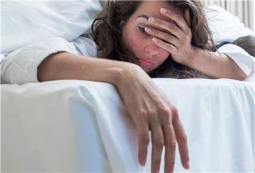 Eine Frau liegt im Bett und fasst sich an den Kopf. Hinter harmlosen Symptomen kann Schlafapnoe stecken.