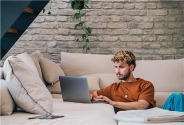 Ein Mann sitzt an seinem Sofa mit einem Laptop. 