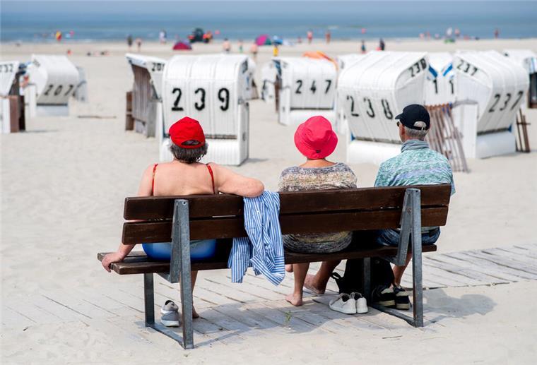 Nordsee statt Mittelmeer: Warum „Coolcation“ zum neuen Reisetrend werden kann Drei Touristen sitzen bei sonnigem Wetter auf einer Bank an einem Strand .