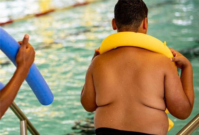Ein übergewichtiger Junge steht mit einer Schwimmnudel am Rande eines Schwimmbeckens. 