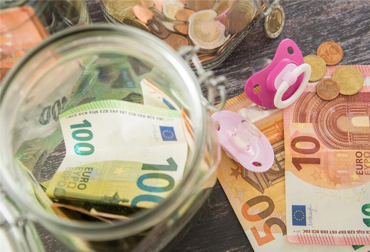 Geldscheine und Münzen sind in gläsernen Behältern und stehen auf einem Tisch. Daneben liegen Schnuller.  Wann landet das Kindergeld auf meinem Konto? Diese Fragen stellen sich jeden Monat viele Familien. Hier gibt es die Termine für die Kindergeld-Auszahlung im August 2025.