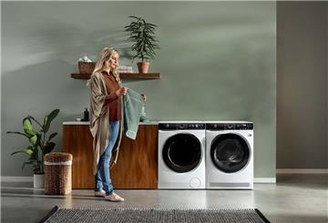 Eine Frau steht in der Nähe von zwei Waschmaschinen und hält ein Kleidungsstück in der Hand. Weiße Schlieren auf dunkler Wäsche entstehen oft durch falsche Dosierung. Moderne Maschinen helfen mit automatischer Dosierung, Rückstände zu vermeiden.
