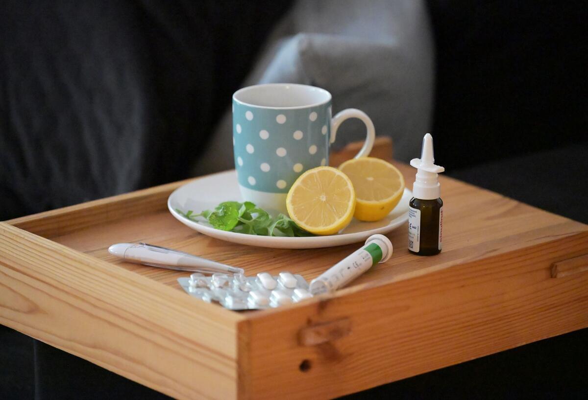 Eine Tasse mit dampfendem Tee samt Zitronenhälften steht auf einem Tablett, daneben befinden sich Nasenspray, Fieberthermometer und Medikamente. (gestellte Szene)