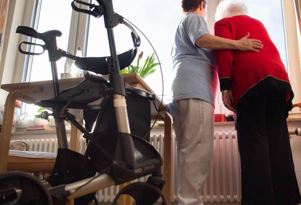 Eine Pflegerin und eine Seniorin stehen am Fenster. 