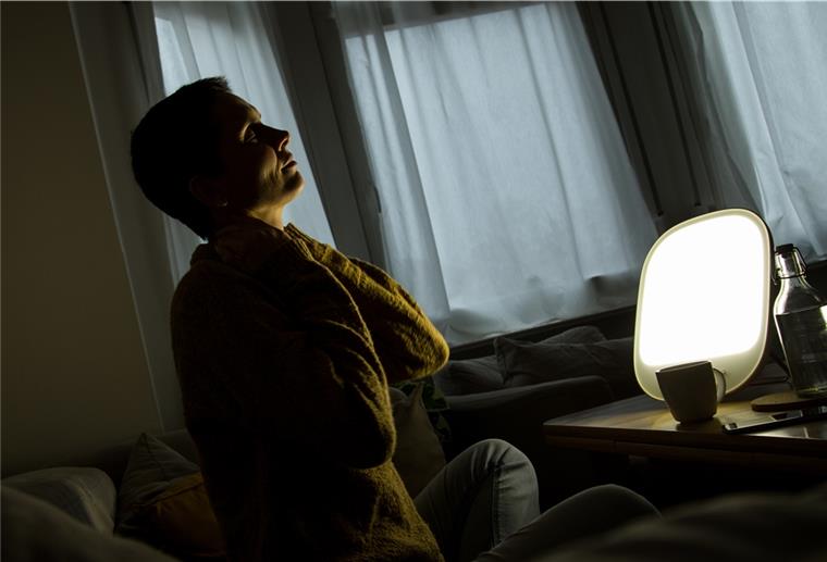 Anwendung von Therapieleuchten braucht Disziplin Eine Frau sitzt in der Dunkelheit vor einer Lampe.