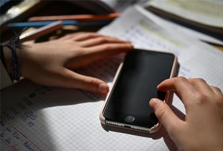 Ein Schulkind sitzt an einem Schreibtisch mit Schulmaterialien und hält ein Smartphone in den Händen. Das Smartphone beeinflusst uns mehr als wir denken. 