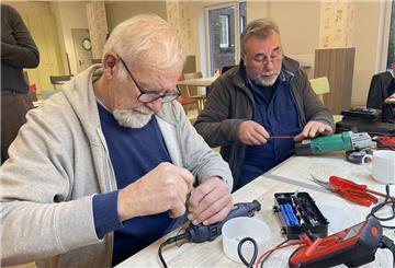 Zwei ältere Männer schrauben an einem Tisch an Elektrogeräten herum.