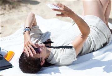 Eine Person liegt am Strand und guckt aufs Handy. Zu viel Sonne schadet dem Handy: Display und Akku sind bei Hitze besonders gefährdet.