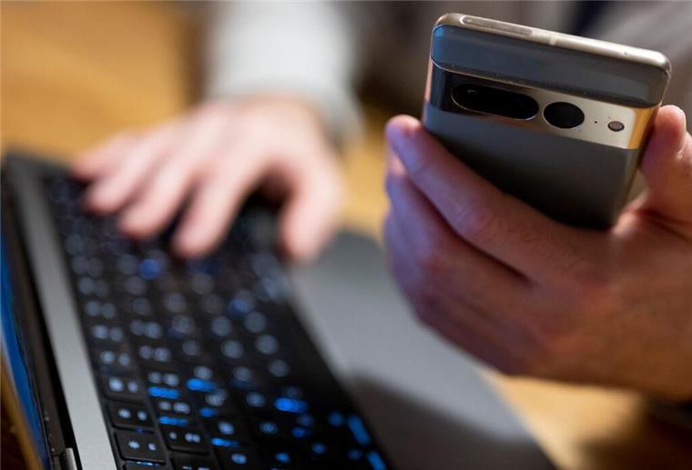 Falsche Beamte am Telefon: So schützt ihr euch vor den Betrügern Ein Mann sitzt am Laptop und hält ein Smartphone mit in der Hand.