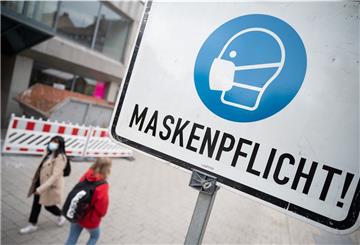 Zwei Passantinnen gehen in einer Fußgängerzone hinter einem Schild vorbei, auf dem „Maskenpflicht“ steht.