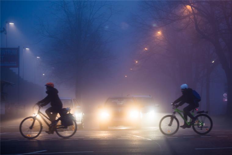 Zwei Radfahrer kreuzen bei Tagesanbruch im Nebel eine Straße. 